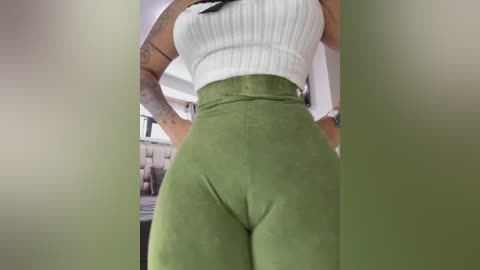 Media: Video of a woman's lower body, wearing a white ribbed crop top and high-waisted, olive-green velvet leggings, showcasing her curvy figure and tattoos on her arms.