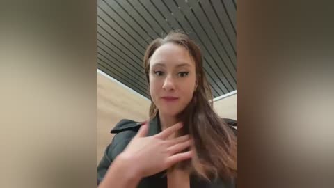 Media: A video of a young woman with light skin and long, straight brown hair, wearing a black leather jacket, standing in an indoor setting with a tiled ceiling.