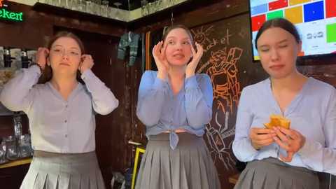 Media: Video of three young women in light blue blouses and gray pleated skirts, hands on their cheeks, in a cozy caf\u00e9 with chalkboard wall art and a TV screen showing a colorful mosaic.