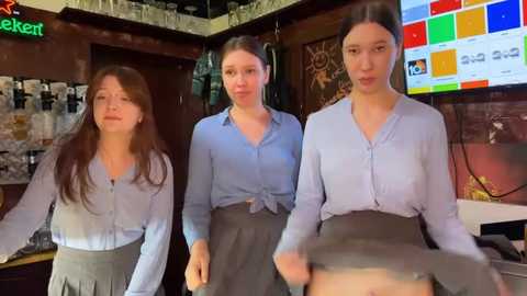 Media: Video of three young women in light blue button-up shirts and gray skirts in a dimly lit bar, with a colorful digital display and bar counter in the background.