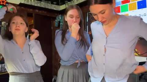 Media: Video of three young women in a bar, one adjusting her hair, another tying a shirt, and the third observing, all in light blue tops and gray skirts. Background includes a colorful mosaic wall, dark wooden decor, and bar shelves.