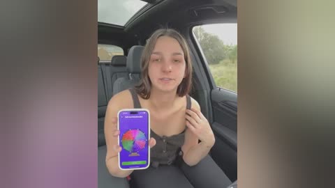 Media: Video of a young woman with shoulder-length brown hair, wearing a black tank top, sitting in a car, holding a smartphone displaying a colorful Pride flag wallpaper.