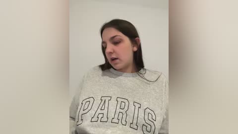 Media: Video of a young woman with long dark hair wearing a gray sweatshirt with \"PARIS\" in bold letters. She stands in a narrow hallway, looking down with a neutral expression. The background is plain and white.