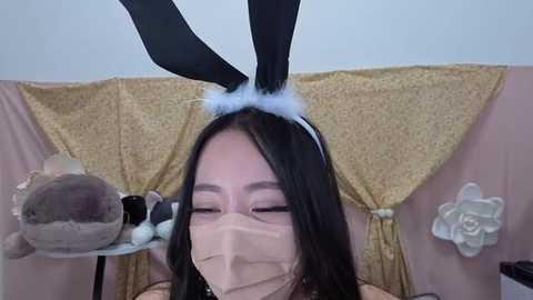 Media: Video of an Asian woman wearing a black bunny ears headband, white mask, and light beige outfit, holding a stuffed animal, with a pink and gold backdrop featuring floral patterns.