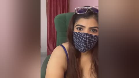 Media: Video of a South Asian woman with long brown hair, wearing a blue checkered face mask, sitting in a green chair, indoors, with a pink wall and red curtains in the background.