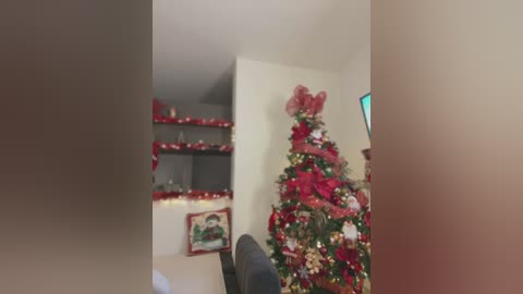 Media: A video of a cozy living room with a decorated Christmas tree adorned with red and gold ornaments, a large red bow, and a red and gold tinsel garland. Shelves above hold festive decorations.