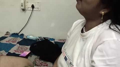 Media: A video shows a woman with dark skin, wearing a white t-shirt and gold earrings, standing in a small room with a blue and red quilt, a black purse, and a pink phone on a bed.