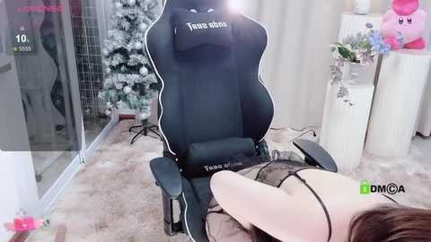 Video of a young woman with long brown hair lying on a plush carpeted floor in a cozy room, wearing black lingerie. She's in a black gaming chair with \"Toshiba\" branding, surrounded by festive decor, including a Christmas tree and plush toys.