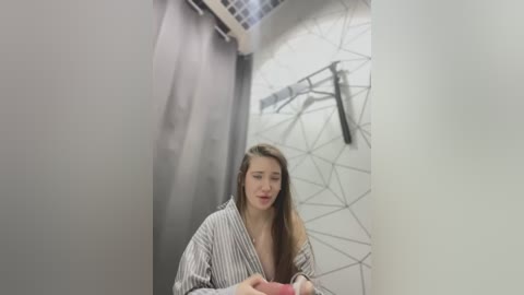 Media: Video of a young woman with long brown hair, wearing a striped robe, sitting in a modern, minimalist bathroom with geometric-patterned walls and a black towel rack.