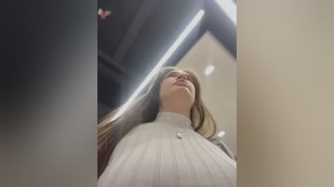 Media: Video of a young woman with long brown hair, wearing a white turtleneck sweater, looking up. Background includes fluorescent lights and a ceiling.