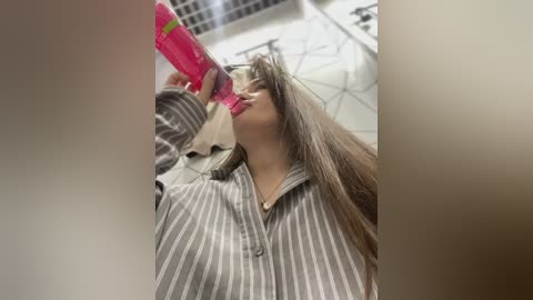 Media: Video of a young woman with long brown hair, wearing a striped pajama top, drinking from a pink water bottle, in a modern room with white walls and a glass staircase.