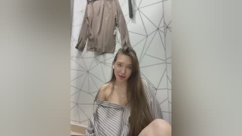 Media: Video of a young woman with long brown hair, wearing a striped off-shoulder top, sitting on a beige floor. Background features a geometric patterned wall and hanging clothes.