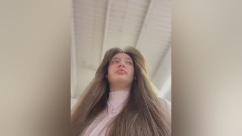 Media: Video of a young woman with long, straight brown hair, wearing a white turtleneck, standing in a brightly lit room with a high ceiling and visible beams.