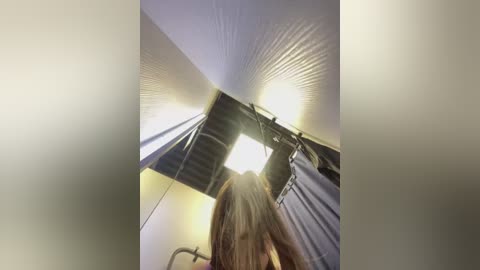 Media: Video of a ceiling with a light fixture, a grey curtain, and a person with long brown hair, viewed from below. The image has a soft focus and is slightly blurred.