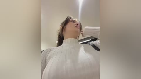 Media: A video of a young woman with fair skin, brown hair, and a white turtleneck sweater, looking upward in a dimly lit room.