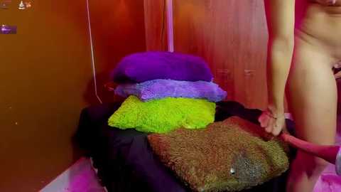 Media: Video of a nude woman with light skin, standing to the right, holding a textured, green blanket. She is partially visible, and a pile of colorful blankets, including purple, green, and black, is on a bed in a warm, reddish room.