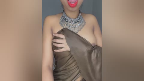 Media: A video of a light-skinned woman with a medium build, wearing a brown, one-shoulder, off-the-shoulder dress with a silver, elaborate necklace. She is playfully sticking out her tongue, and her hand is modestly covering her breast.