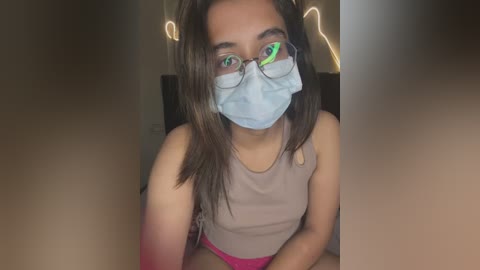 Media: Video of a young woman with light brown skin and long brown hair, wearing a face mask, glasses, and a sleeveless top, sitting in a dimly lit room with string lights in the background.