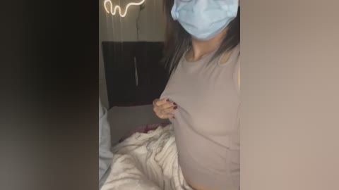 Media: Video of a pregnant woman with a blue mask, light brown top, and dark hair, adjusting her bra strap, in a dimly lit bedroom with a neon light and bed.