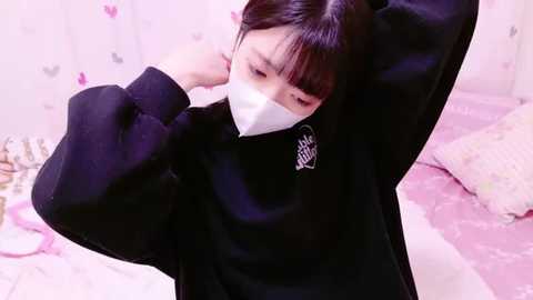 Media: Video of an East Asian woman with straight black hair and a white mask, adjusting a black hoodie in a pastel-colored, butterfly-patterned room.