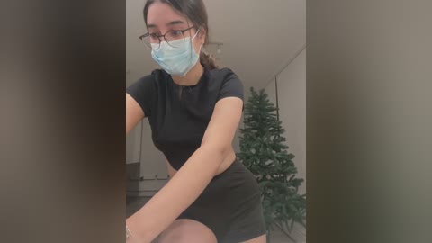 Video of a young woman with fair skin, wearing glasses, a blue surgical mask, and a black dress, posing indoors beside a Christmas tree.