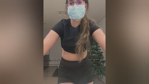 Video of a woman in a black crop top and shorts, wearing a blue face mask and glasses, standing indoors with a green plant and white walls in the background.