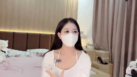 Media: Video of a young Asian woman with long black hair, wearing a white face mask and top, standing in a modern bedroom with beige walls, a brown upholstered headboard, and light-colored curtains.