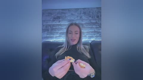 Media: Video of a blonde woman with fair skin, seated in a dark leather chair, holding a plate of fries. Background features a white brick wall under dim lighting.