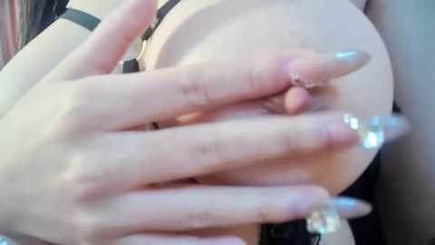 Media: Video of a light-skinned woman's hands and chest. Her manicured nails are long with glittery silver polish. She is wearing a black bra and has a tattoo on her right wrist.