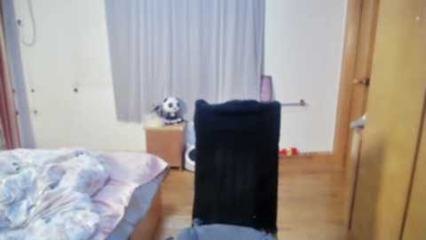 Media: Video of a cluttered bedroom with a bed, a black chair, a stuffed animal, and a closed door; beige walls and wooden floor.