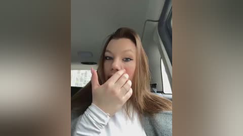 Media: Video of a young Caucasian woman with light skin and long blonde hair, wearing a white shirt, holding her mouth with her hand, sitting in a car.