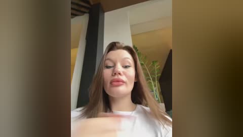 Media: A video of a young Caucasian woman with fair skin and straight brown hair, wearing a white T-shirt, sitting indoors near a window with a green plant in the background.