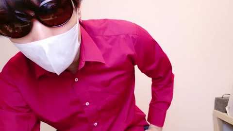 Media: Video of a person in a red button-down shirt and white face mask, wearing sunglasses, with hands in pockets, against a plain beige background.