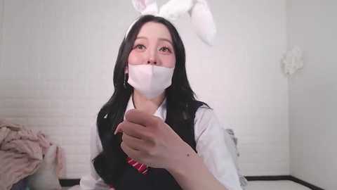 Media: A video of an Asian woman with long black hair, wearing a white bunny costume and a surgical mask, in a white-tiled room, holding a glass.