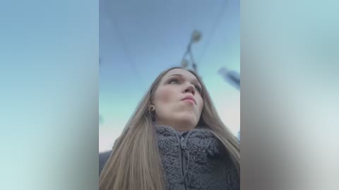 Media: A video captures a young woman with long blonde hair, wearing a gray fleece jacket, looking up at a clear blue sky with a blurred lamp post in the background.