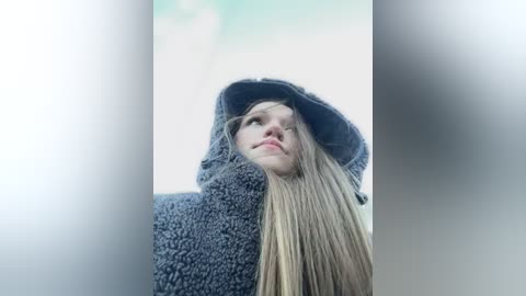 Media: Video of a young blonde woman with long hair, wearing a gray hooded sweatshirt, looking up thoughtfully against a soft-focus, cloudy sky background.