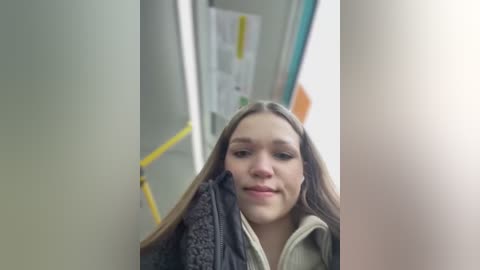 Media: A video of a young woman with long brown hair, wearing a grey fleece jacket, standing in a narrow hallway with yellow handrails. The background features posters and a light-colored wall.