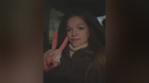 Media: A dimly lit video of a young woman with long brown hair, wearing a black hoodie and white shirt, making a peace sign with red-painted nails, standing in a dark, possibly urban, setting.