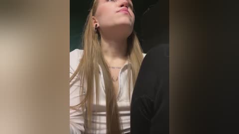 Media: Video of a young woman with long blonde hair, wearing a white blouse, gazing upward with a serene expression. The background is blurred, with a hint of a dark figure on the right.