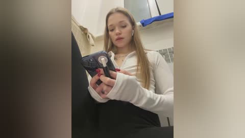 Media: Video of a young woman with long blonde hair, wearing a white hoodie, sitting on a black car seat, playing with a small pink toy. The background shows a beige bathroom with a blue towel.