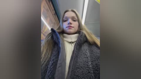 Media: A blurry video of a young woman with long blonde hair, wearing a dark grey puffer jacket over a cream turtleneck, standing in a modern indoor setting with fluorescent lighting.