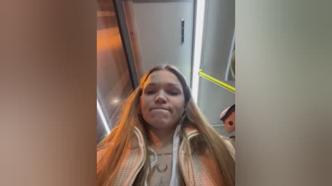 Media: Video of a young Caucasian woman with long blonde hair, wearing a beige jacket, standing in a subway car. She has a neutral expression. The background shows a yellow railing and gray walls.