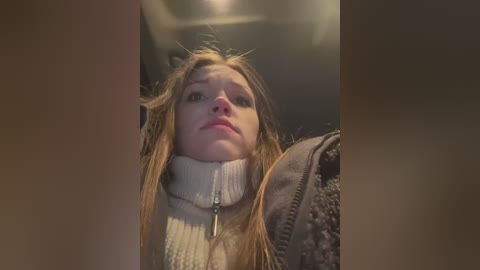 Media: A video captures a young, fair-skinned woman with long, messy blonde hair, wearing a cream-colored turtleneck sweater, looking distressed with wide eyes and a furrowed brow. She stands in a dimly lit, cluttered room with a blurred background.