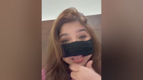 A video of an Asian woman with long, light brown hair, wearing a black face mask, giving a thumbs-up gesture. The background is a beige wall.