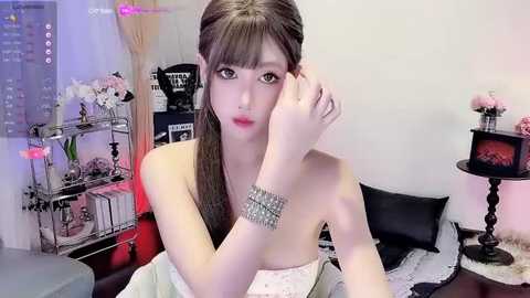 Media: Video of a young Asian woman with long, straight brown hair, fair skin, and delicate features, sitting on a bed, wearing a white top, silver bracelet, and makeup, in a modern, minimalistic room with a cityscape view.