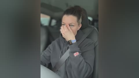 Media: Video of a woman with light skin, dark hair tied back, wearing a gray hoodie and seatbelt, covering her face with her hand, sitting in a car.