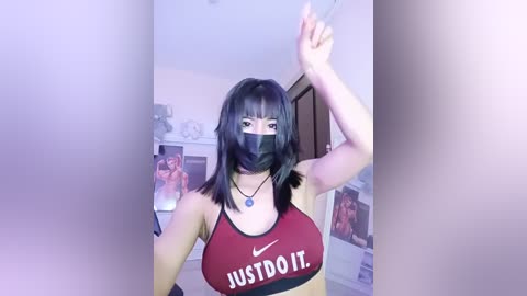 Video of a young Asian woman with straight black hair, wearing a black face mask, red Nike sports bra, and a black choker. She poses indoors with a blurred background.