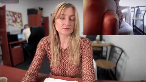 Media: Video of a blonde, Caucasian woman with long hair, wearing a red and white patterned blouse, sitting at a desk in an office. Background includes a bulletin board, bookshelves, and a chair.