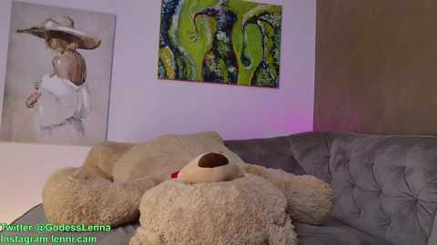 Media: Video of a plush teddy bear on a gray couch in a room with a large, colorful abstract painting on the wall. Text at the bottom shows social media handles.