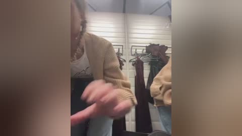 Media: Video of a young woman with light skin and curly brown hair, wearing a beige sweater, standing in a store with a mirror reflecting her reflection and hanging clothes.